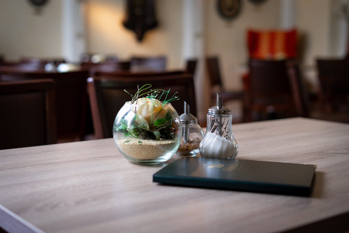 Gemütlicher Tisch im Café mit einer runden Glasvase inklusive Sand und Blumengesteck, Zuckerdosen und einer geschlossenen Speisekarte im Vordergrund.