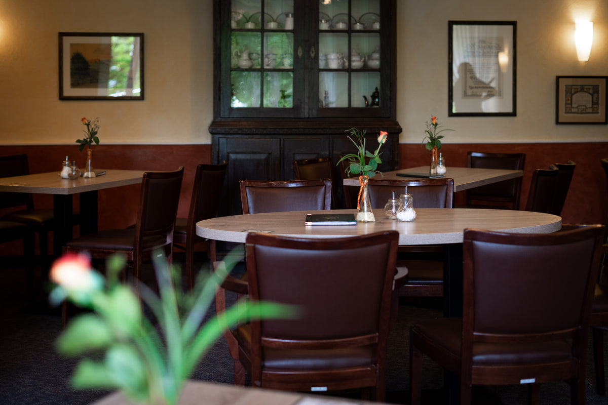 Gemütlicher Blick in den warmen Innenraum des Cafés mit mehreren eingedeckten Holztischen, dunklen Lederstühlen und einer großen Vitrine im Hintergrund.