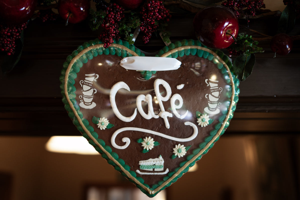 Lebkuchenherz in Schutzfolie mit der weißen Aufschrift "Café", verziert mit grünem Zuckerguss, kleinen Blümchen und Kaffeesymbolen, aufgehängt unter einer Dekoration aus roten Äpfeln und Beeren.