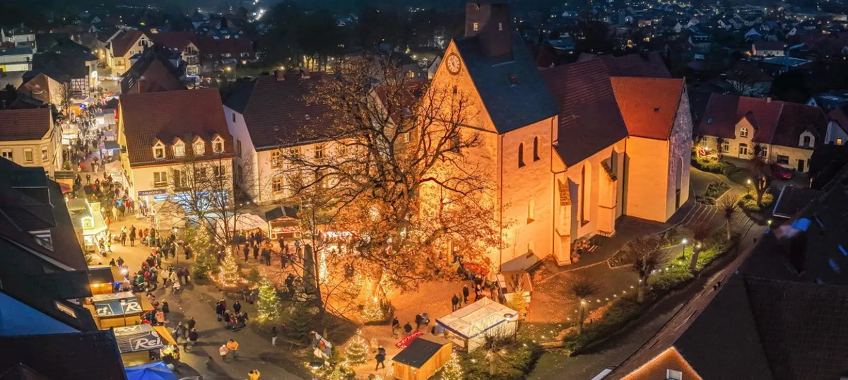 Luftaufnahme des stimmungsvollen Weihnachtsmarktes in Borgholzhausen bei Nacht. Die historische Kirche ist festlich beleuchtet und bildet das Zentrum, umgeben von zahlreichen geschmückten Marktständen und Besuchern.