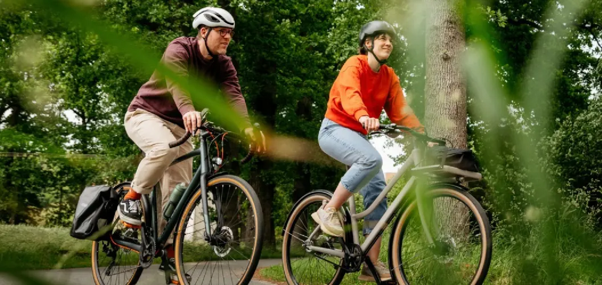 Zwei Personen mit Fahrradhelmen fahren auf Fahrrädern durch eine grüne Natur- und Parklandschaft, im Vordergrund teilweise von Blättern und Zweigen verdeckt.