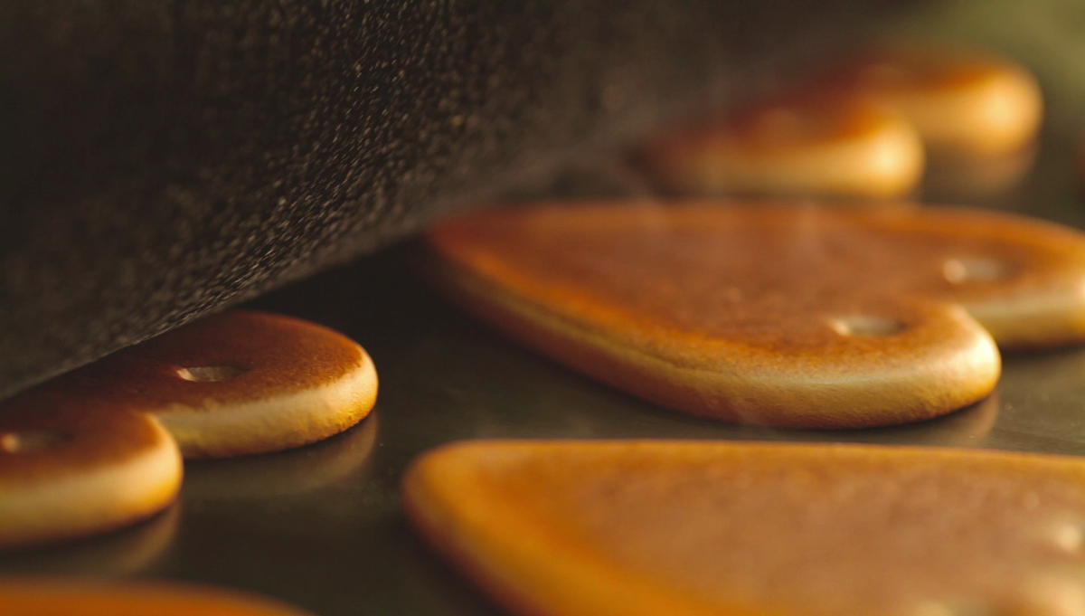 Nahaufnahme aus der Produktion von Lebkuchenherzen: Eine dunkle, texturierte Formwalze rollt über die Teiglinge, die auf einer glatten Produktionsfläche liegen. Die Lebkuchenherzen sind bereits ausgestanzt und weisen jeweils kleine, für die Aufhängung typische Löcher auf.