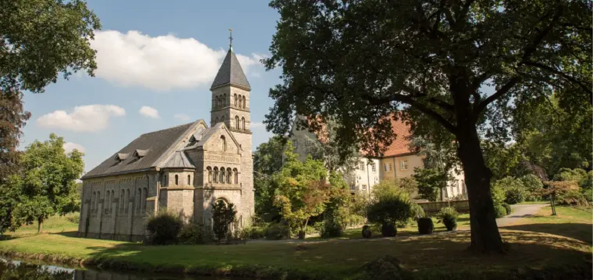 Ansicht von Schloss Brincke entlang der Radroute R10, mit der Schlosskapelle im Vordergrund und dem Hauptgebäude im Hintergrund, umgeben von einem parkähnlichen Gelände mit altem Baumbestand.
