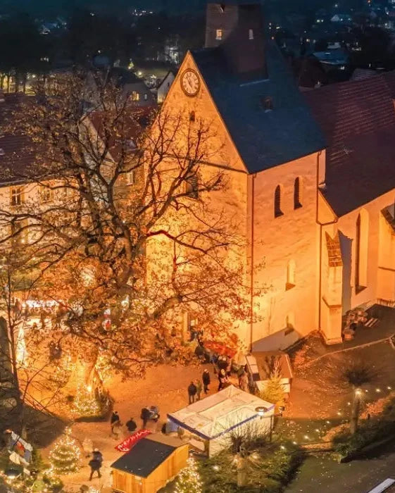 Luftaufnahme des stimmungsvollen Weihnachtsmarktes in Borgholzhausen bei Nacht. Die historische Kirche ist festlich beleuchtet und bildet das Zentrum, umgeben von zahlreichen geschmückten Marktständen und Besuchern.