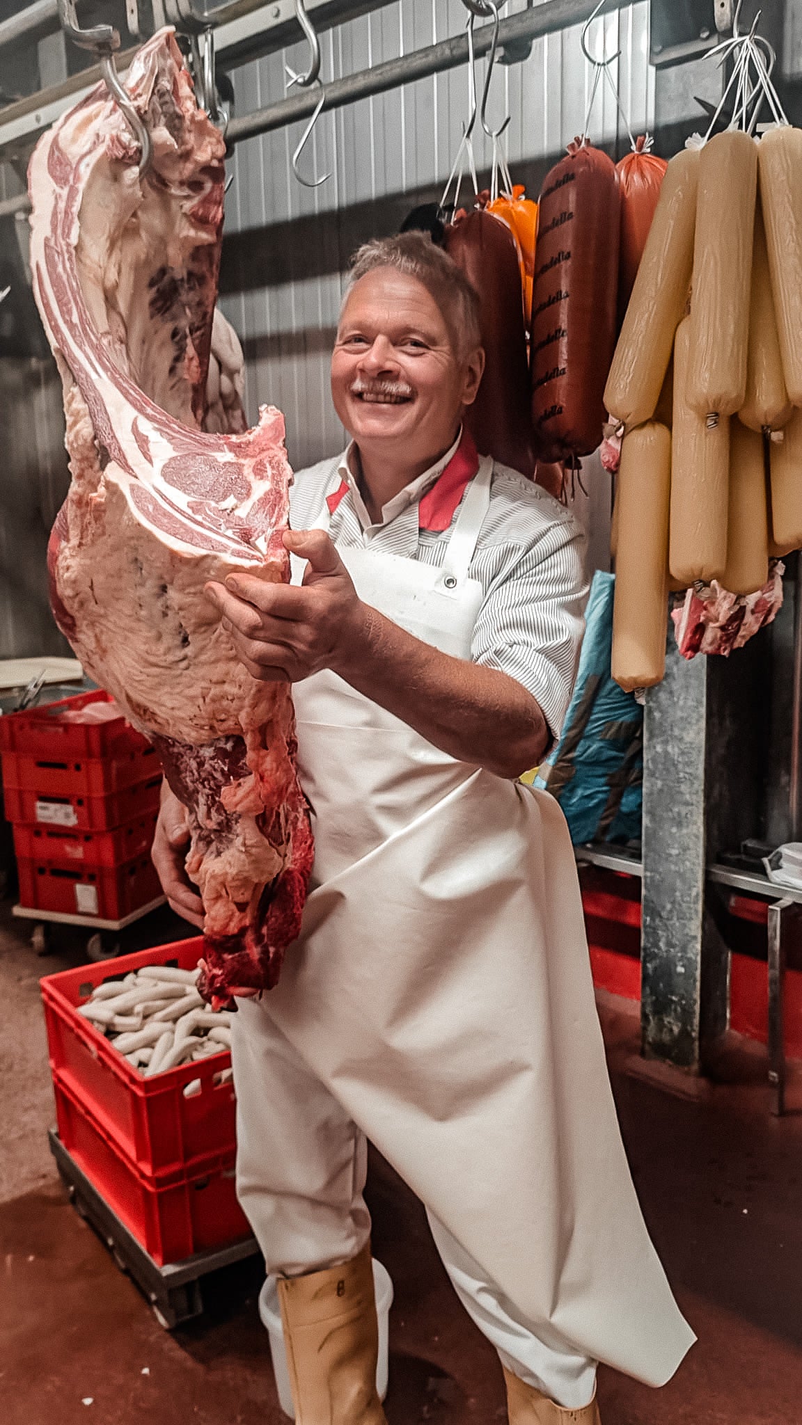 Ein gut gelaunter Fleischer in weißer Schürze präsentiert in der Fleischerei Goldbecker stolz ein großes Stück frisches Fleisch, während im Hintergrund Wurstwaren hängen.