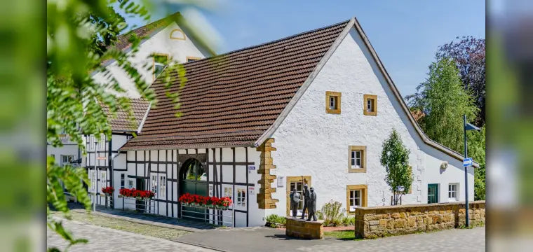 Ansicht des historischen Kultur- und Heimathauses in Borgholzhausen, einem Gebäude mit weiß verputzten Wänden, traditionellem Fachwerk und einem großen Satteldach. Im Vordergrund sind kleine Bronzefiguren und eine niedrige Steinmauer zu sehen.