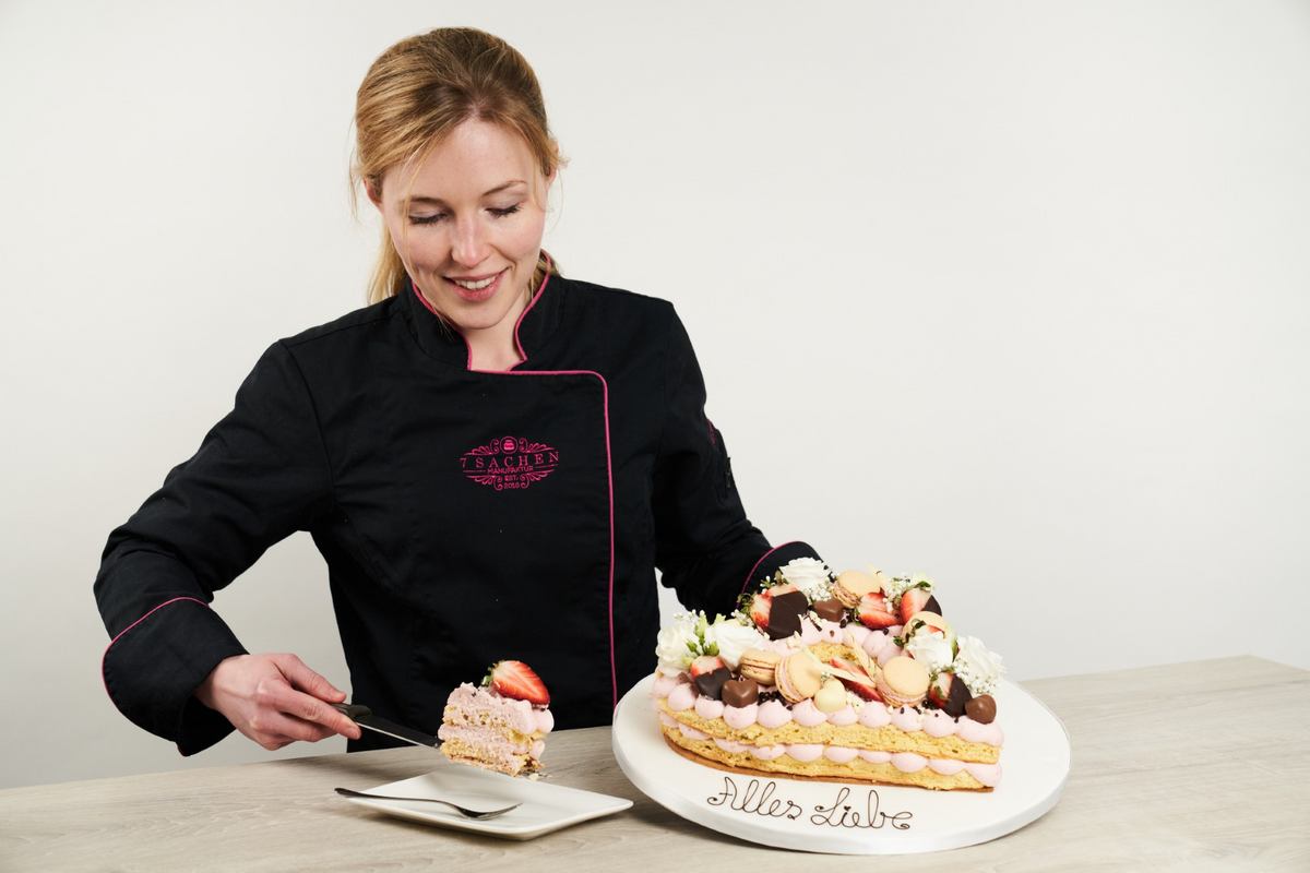 Eine lächelnde Konditorin in schwarzer Uniform mit rosa Rand und Logo serviert ein Stück einer reich verzierten herzförmigen Torte auf einem weißen Teller mit der Aufschrift "Alles Liebe".